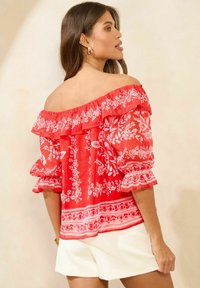 Rød off-shoulder bluse med hvide blomster mønstre, flæseærmer og elastisk halsudskæring; parret med hvide shorts.