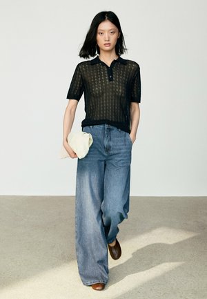 Donna che cammina indossando una polo nera semitrasparente in maglia, jeans blu a gamba larga, scarpe marroni e che tiene in una mano un panno beige piegato.