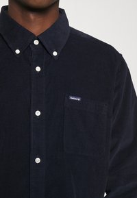 Homme portant une chemise en velours côtelé bleu foncé avec des boutons blancs et une poche poitrine étiquetée Barbour.