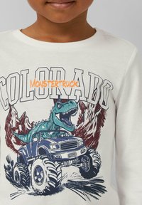 Weißes Langarmshirt mit einem blauen Monstertruck und einem grünen Dinosaurier vor roten Bergen, mit dem Text "COLORADO" und "MONSTERTRUCK".