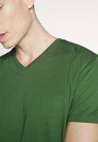 Närbild av en person som bär en enkel grön V-ringad t-shirt, synlig nacke, axel och del av ansikte mot en vit bakgrund.