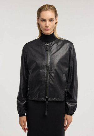 Frau mit nach hinten gegelten blonden Haaren, die eine schwarze Lederjacke mit Reißverschluss über einem schwarzen, hochgeschlossenen Oberteil vor einem einfarbigen hellen Hintergrund trägt.