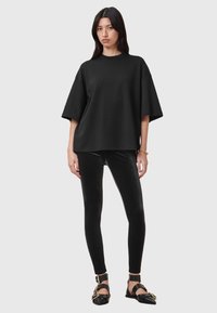 Donna che indossa una t-shirt nera oversized, leggings in velluto nero, ballerine nere con fibbie dorate e un bracciale dorato, in piedi su uno sfondo grigio.