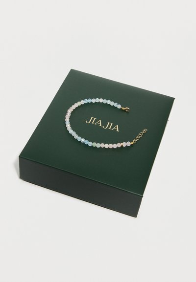 Bracelet perlé avec des pierres facettées de couleur pastel et une fermeture en or, posé sur une boîte vert foncé avec le texte doré "JIAJIA".