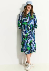 Blumenkleid mit blauen, grünen und schwarzen Mustern; Knopfleiste, Puffärmel, gestuftem Rock. Kombiniert mit weißen Sneakern und einem blauen Bucket Hat.