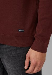 Bordowy sweter z dzianiny z żebrowanymi mankietami i dołem, z małą niebieską metką "Elevated Casual" na dole.