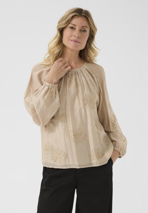 Blouse beige brodée à manches longues, col froncé, tissu texturé, motifs floraux et poignets élastiques. Portée avec un pantalon noir.