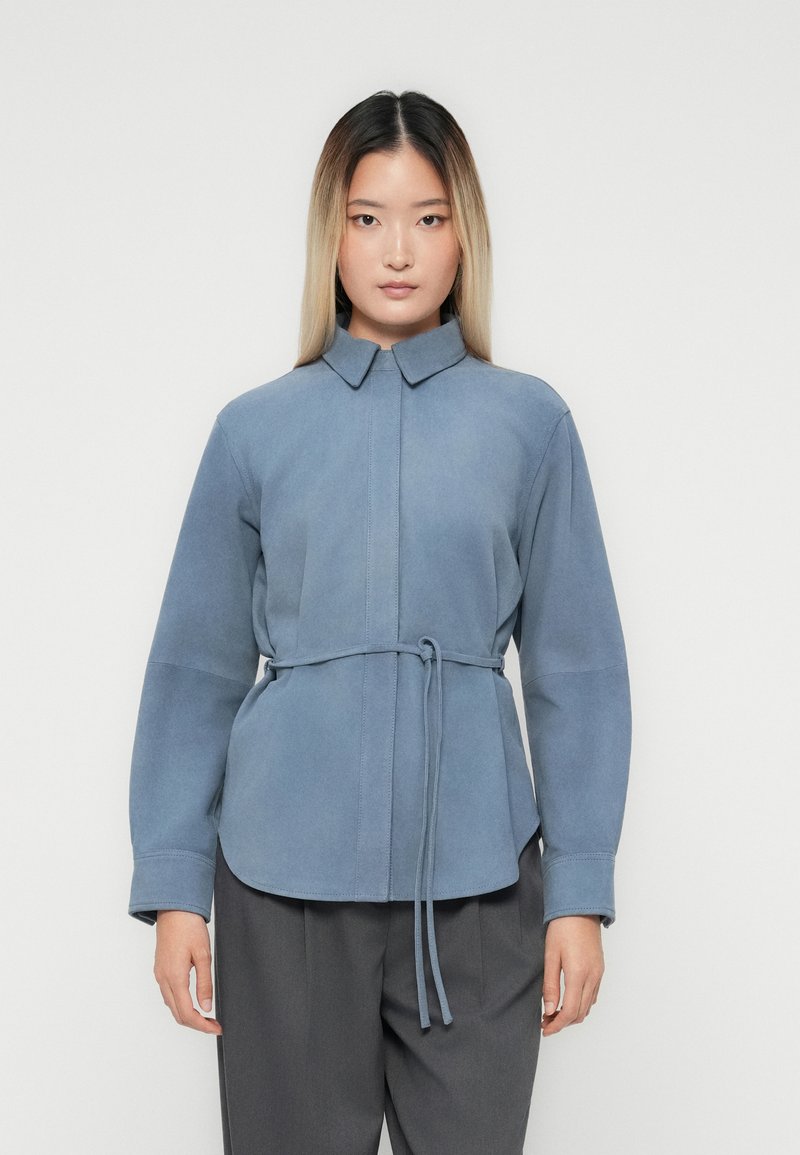 Vrouw met steil ombre haar, gekleed in een blauwe suede jas met riem en grijze broek, staande tegen een effen lichte achtergrond.