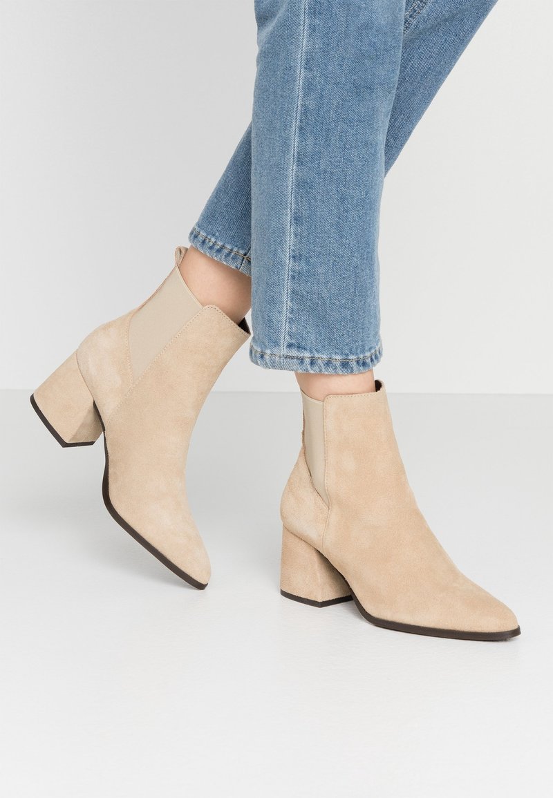 Bottines en suède beige avec un bout pointu, des panneaux latéraux élastiques et un talon épais. Surface texturée et lignes épurées.