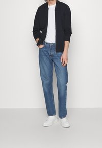 Adulto vestido con una chaqueta negra con cremallera sobre una camiseta blanca, jeans azules y zapatillas blancas, de pie con una mano en el bolsillo.