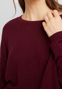 Burgundy- värinen neule, jossa on pyöreä pääntie, ribbaatut hihansuut ja pehmeä rakenne. Läheltä otettu kuva, joka korostaa kangasta ja yksityiskohtia hihassa.