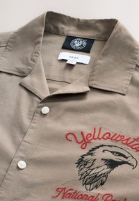 Chemise marron clair à manches courtes avec col, dotée de boutons blancs et d'un texte brodé rouge ainsi que d'un dessin d'aigle, étiquetée "Yellowstone National Park".