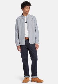 Camisa abotonada gris claro sobre una camiseta blanca, jeans oscuros y botas marrones. La camisa presenta un cuello redondeado y un detalle de logo.