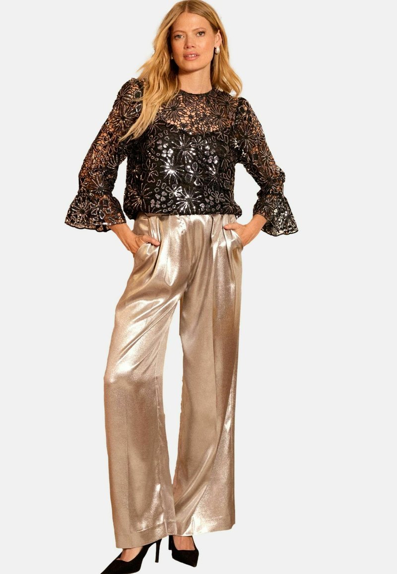 Top in pizzo floreale nero con maniche a campana; pantaloni larghi metallici dorati; modella con le mani nelle tasche; tacchi neri.