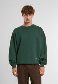 Zelená oversize mikina vyrobená z mäkkého materiálu, s okrúhlym výstrihom a manžetovými rukávmi. Biele logo na dolnom ľavom rohu.