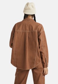 Camicia oversize marrone con cuciture bianche a contrasto, dotata di orlo curvo, polsini con bottone e una vestibilità comoda. Indossata con pantaloni abbinati.