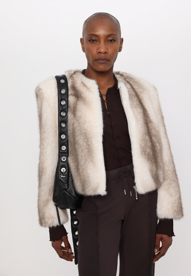 Chaqueta corta de piel sintética en color beige con una correa de cuero negro que tiene broches metálicos; combinada con una blusa marrón oscura y pantalones texturizados.