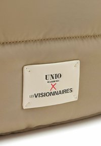 Borsa beige imbottita con tessuto testurizzato. Presenta un'etichetta in pelle bianca con "UNIO HAMBURG X LES VISIONNAIRES" in testo nero e rosso.