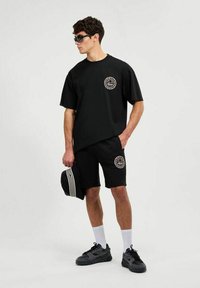 Conjunto de camiseta de algodón negro y pantalones cortos con logo en el pecho, combinado con una gorra negra y zapatillas negras. El modelo lleva gafas de sol oscuras.