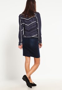 Marinblå tröja med geometriska mönster och vita detaljer, kombinerad med en mörk denimpencilkjol och svarta ballerina, sedd bakifrån.