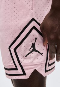 Κοντινό πλάνο χεριού με σκούρο δέρμα που ακουμπάει κοντά στο στρίφωμα ροζ σορτς μπάσκετ με μαύρες τρύπες και μαύρο λογότυπο Jumpman.