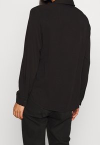 Chemise noire à manches longues avec poignet boutonné et ourlet légèrement incurvé. Tissu doux à texture lisse, présentant des lignes épurées et un design minimaliste.