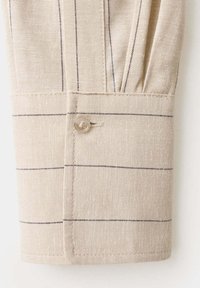 Poignet de chemise en tissu beige avec de fines rayures noires verticales et horizontales, sécurisé par un petit bouton translucide.
