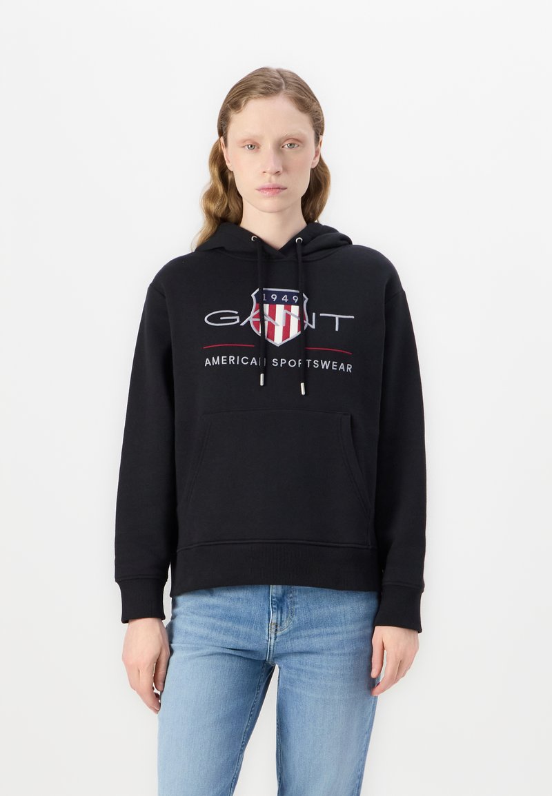 Black hoodie made of cotton, featuring a front pocket, drawstring hood, and red/white logo design. Worn with light blue jeans.