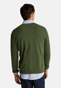 Mężczyzna widoczny od tyłu, ubrany w oliwkową bluzę na jasnoniebieską koszulę i ciemne dżinsy, na tle jednolitego białego tła.