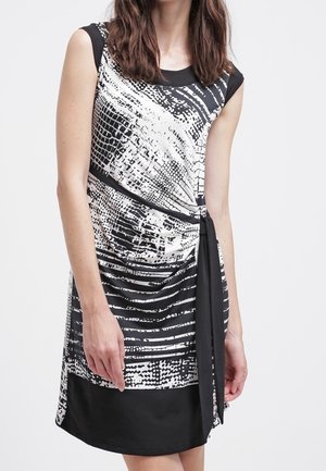 Frau trägt ein ärmelloses schwarz-weiß gemustertes Kleid mit schwarzem Seitendetail, stehendes vor einem schlichten hellen Hintergrund.