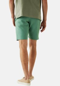 Pantaloni corti in cotone verde menta con vestibilità rilassata, dotati di tasche laterali e una texture liscia. Indossati con una maglia verde oliva a taglio largo.