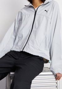 Jasnoszara kurtka zapinana na zamek z wysokim kołnierzem, czarnym zamkiem błyskawicznym i logo. Posiada luźne rękawy i elastyczne mankiety. Noszona na czarnych spodniach.