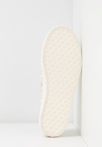 Suola di sneaker bianca con un motivo a trama esagonale, posizionata verticalmente contro un angolo bianco di parete e pavimento.