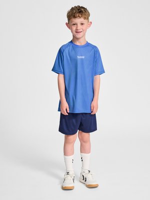Junge mit lockigem Haar, der ein blaues Sport-T-Shirt, dunkelblaue Shorts, weiße Socken und weiße Turnschuhe trägt, steht und lächelt.