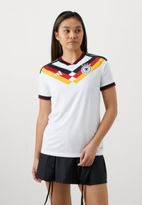 Femme portant un maillot de soccer blanc avec un motif chevron rouge, jaune et noir, et une jupe de sport noire sur un fond uni.
