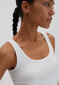 Witte tanktop gemaakt van een zachte katoenkwaliteit met brede banden en een ronde hals. De afbeelding toont een zijprofiel en textuurdetails.