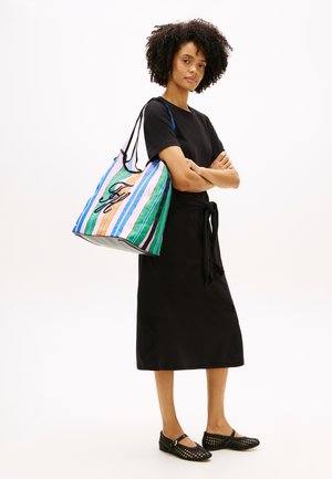 Frau mit lockigem Haar in einem schwarzen Kleid und schwarzen gewebten Ballerinas, die eine große bunte, gestreifte Tragetasche über der Schulter trägt und die Arme verschränkt hat.