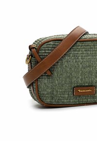 Bolso cruzado de tela verde trenzada con detalles y correa de cuero marrón, que cuenta con un cierre de cremallera y un parche con el logotipo en la parte frontal.