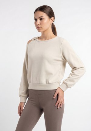 Ung kvinde med brunt hår i hestehale har beige sweatshirt med udskæring på skulderen og brune, tætsiddende leggings og står med den ene hånd på hoften.