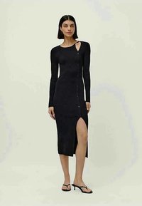 Mulher vestindo um vestido midi preto de malha com uma fenda até a coxa, corte nos ombros, mangas longas e detalhes em botão, combinado com sandálias pretas de salto.