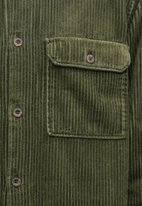 Μπλούζα γουτουρέ σε ελαιόλαδο πράσινο με κάθετες ραβδώσεις, τσέπη στο στήθος και καφέ κουμπιά για κλείσιμο. Μαλακή υφή με δομημένο σχέδιο.