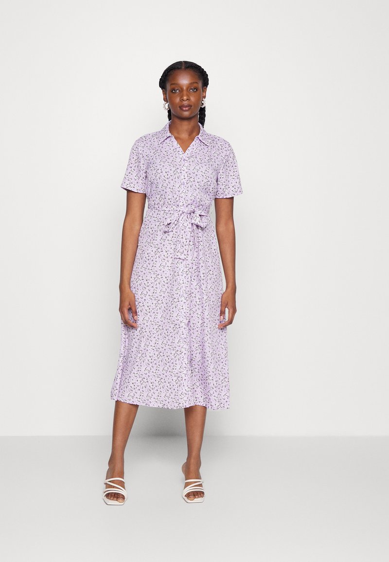 Vestido floral roxo com mangas curtas, abertura frontal com botões, cintura ajustável e comprimento até ao meio da perna. Apresenta pequenos padrões de flores espalhadas por todo o tecido.