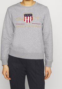 Grå sweatshirt med rund hals, der har et broderet skjoldlogo i rødt og guld samt tekst. Ribstrikkede manchetter og kant med en blød tekstur.