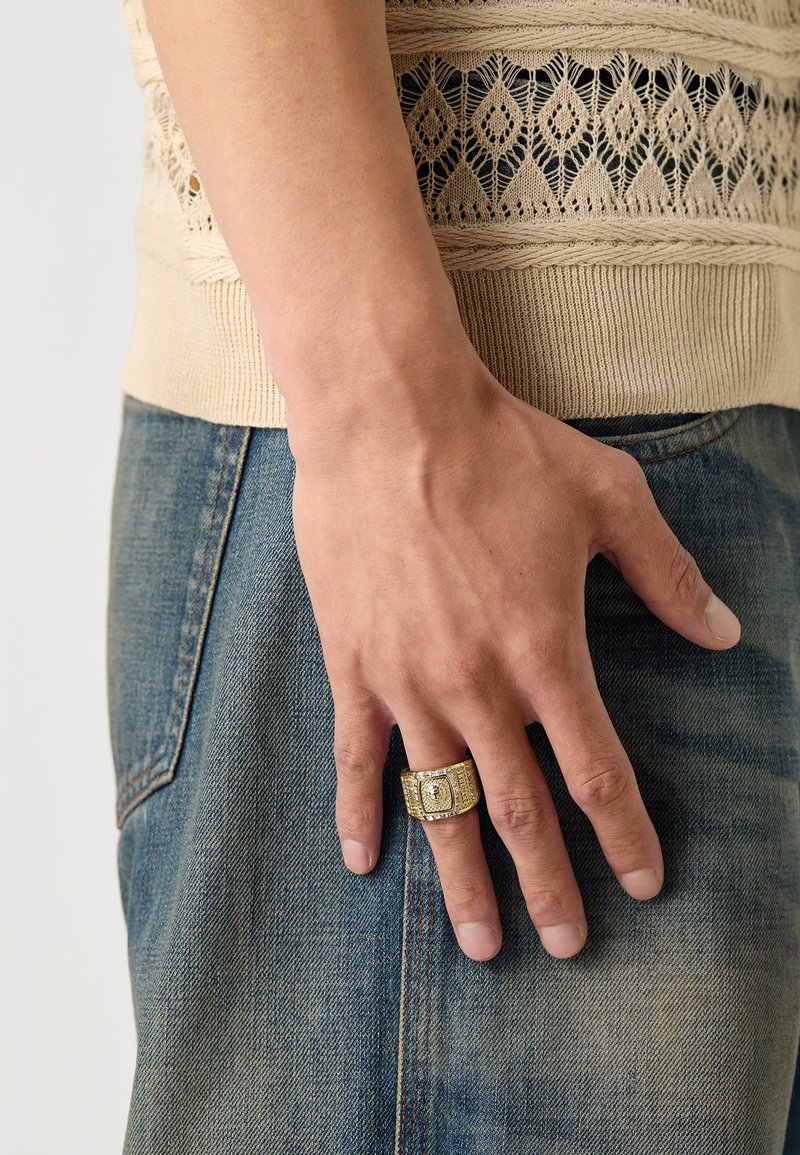 Kuldne sõrmus, millel on ristküliku kujuline näo ja selge kivi, kantud käel, mis resting blue denim kangal. Kreemjas kudum, millel on tekstuurilised detailid.