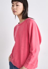 Sudadera rosa de corte relajado con mangas raglán y detalle bordado en el puño izquierdo. Tejido suave y texturizado con un diseño casual.