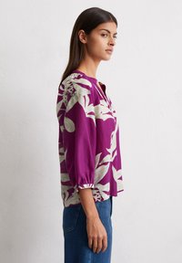 Chemisier violet avec de grands motifs floraux blancs, présentant un col rond et des manches trois-quarts froncées aux poignets.