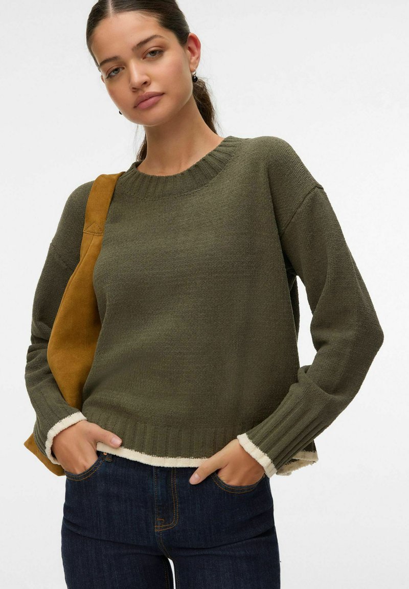 Suéter de punto verde oliva con un cuello redondo acanalado y dobladillo blanco en contraste. Mangas largas y corte relajado. Combinado con jeans oscuros.