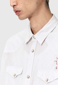 Λευκό πουκάμισο με ρίγες και γιακά με κουμπιά, διαθέτει δύο εμπρόσθιες τσέπες, μεταλλικά κουμπιά ως διακοσμητικά στοιχεία και κόκκινη κεντητή ανθοδέσμη στο στήθος.