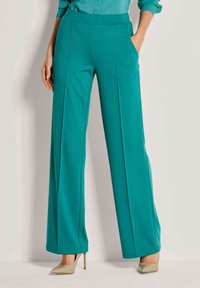 Vrouw in teal hoge taille wijde broek en beige puntige hakken, staande met één hand in de zak tegen een effen achtergrond.