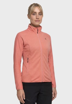 Frau trägt eine korallrosa Jacke mit Reißverschluss und hohem Kragen sowie schwarze Hose, steht vor einem einfarbigen hellen Hintergrund.